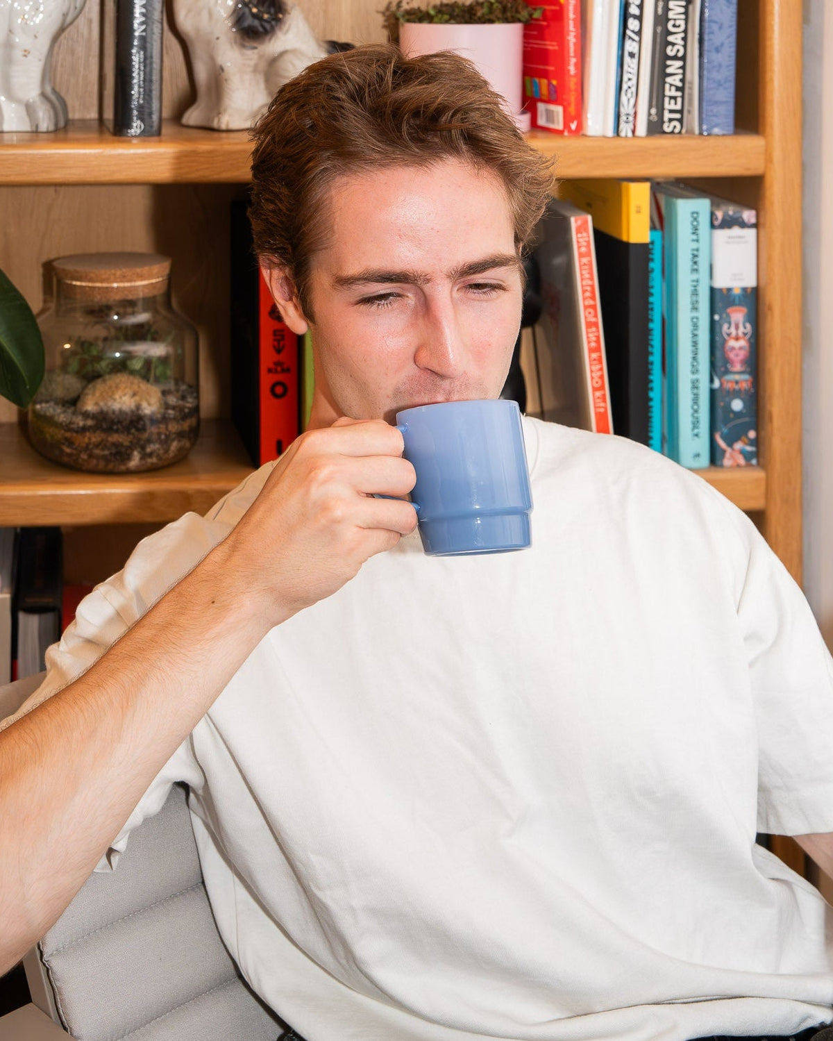 A man in a white t-shirt sitting in a chair in front of a wooden bookshelf, drinking from a light blue ceramic mug.